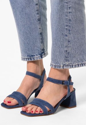 Blue suede heeled sandals with two wide straps across the toes, ankle straps, and a square block heel, worn with cropped denim.