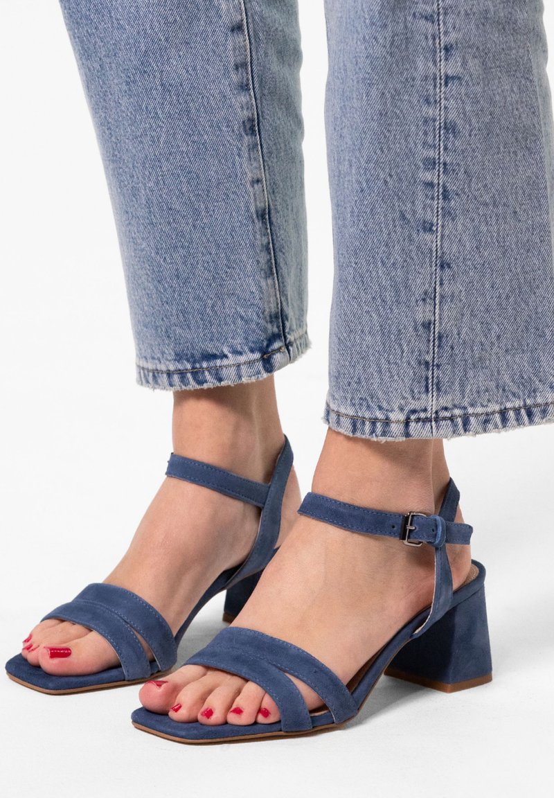 Blaue Wildleder-Absatzsandalen mit zwei breiten Riemen über den Zehen, Knöchelriemen und einem quadratischen Blockabsatz, getragen mit einer verkürzten Jeans.