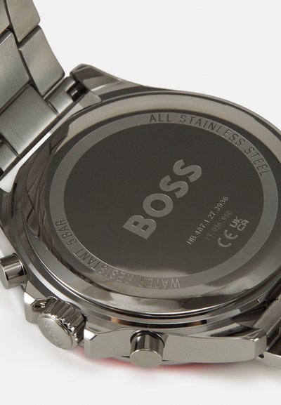 Fond de montre en acier inoxydable avec logo et texte gravés. Présente un boîtier circulaire lisse, une couronne proéminente et une finition métallique brossée.