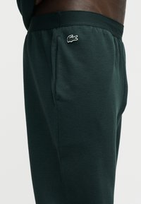 Gros plan sur un pantalon texturé vert foncé avec une poche latérale et un petit logo crocodile blanc Lacoste sur la hanche, porté par une personne.