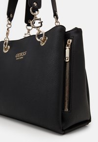 Mustan tekstuuripintaisen nahkaisen Guess-laukun kultaiset yksityiskohdat, logo-riipus hihnassa, etupuolen logo-teksti ja vetoketjutasku sivussa.