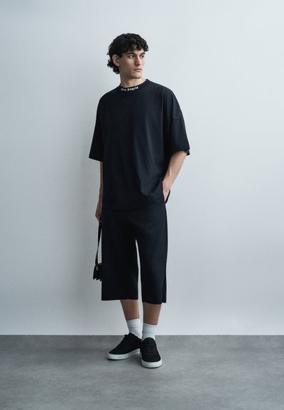 Tânăr purtând un tricou negru supradimensionat, pantaloni scurți largi negri, șosete albe, adidași negri, ținând o geantă mică neagră, stând în fața unui perete gri.