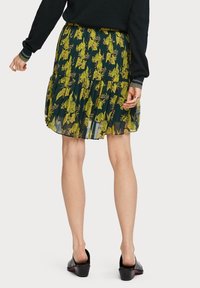 Jupe plissée florale en vert foncé avec des motifs jaunes, dotée d'une taille élastique et d'un ourlet superposé. Associée à des chaussures noires à enfiler.