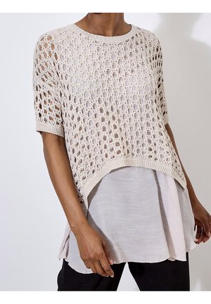 Personne portant un haut beige en maille ajourée à manches courtes superposé sur un chemisier léger et ample avec un pantalon noir.
