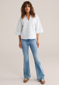 White pinstriped button-up shirt with wide sleeves, paired with light blue flared jeans and brown sneakers, standing against a neutral background.