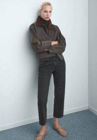 Mujer con cabello rubio apoya su espalda en la pared, vestida con una chaqueta marrón con cuello, jeans negros cortos y zapatos planos beige sobre una alfombra azul.