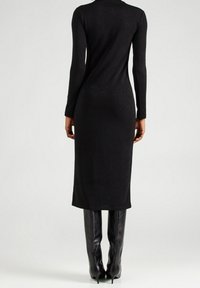 Eine Person trägt ein enganliegendes, langärmliges schwarzes Midikleid, kombiniert mit knielangen schwarzen Lederstiefeln mit Absatz, von hinten gezeigt.