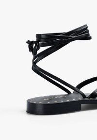 Sandalo in pelle nera con cinturini incrociati e una decorazione annodata. La suola piatta presenta motivi circolari in oro e bianco su uno sfondo nero.