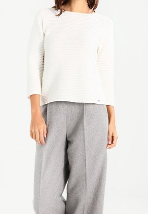 Vrouw draagt een wit geribd shirt met lange mouwen en losse grijze wollen broek, staand tegen een effen witte achtergrond.