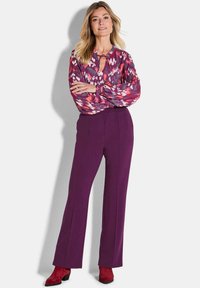 Langärmlige Bluse mit einem lebendigen pinken und lila Muster, ausgestattet mit einem Schlüssel-Ausschnitt. Kombiniert mit hohen lila Hosen und roten, hochhackigen Stiefeln.