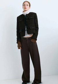 Czarna teksturowana krótka kurtka z dwoma przednimi kieszeniami, zapinana na guziki, noszona na jasnym golfie, w połączeniu z brązowymi spodniami o szerokich nogawkach.