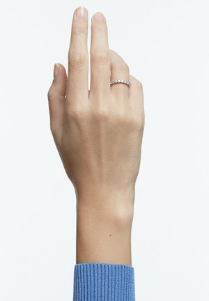 Damska ręka z srebrnym pierścionkiem zdobionym małymi diamentami na palcu serdecznym, na białym tle, ubrana w niebieski prążkowany rękaw.