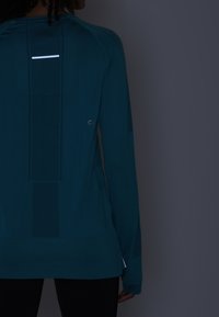 Langärmliges Sportoberteil in Petrolblau mit atmungsaktiven Mesh-Einsätzen, reflektierenden Rückendetails und einem lockeren Schnitt mit Seitenschlitzen.