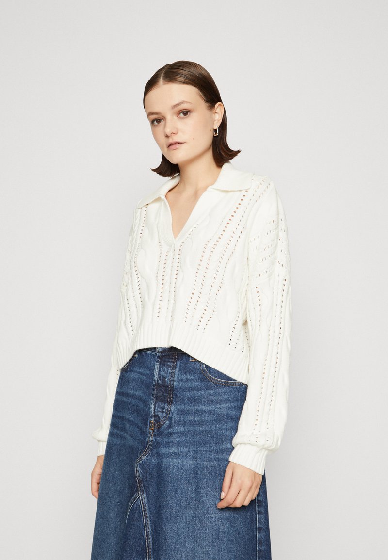 Maglione bianco lavorato a maglia con colletto, caratterizzato da motivi a treccia e dettagli in pizzo, abbinato a una gonna in denim scuro.