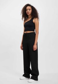 Czarny crop top na jedno ramię w zestawieniu z luźnymi czarnymi spodniami z kieszeniami i sznurkiem w pasie oraz białymi sneakersami. Gładka faktura materiału.