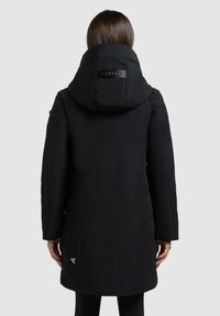 Zwarte geïsoleerde parka met een grote capuchon. Bevat een logo-detail op de nek en een klein merkembleem dicht bij de zoom. Gladde, matte textuur.