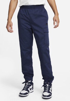 Mann trägt marineblaue Cargo-Jogginghosen mit elastischen Bündchen und schwarz-weißen High-Top-Sneakern, steht vor einem weißen Hintergrund.