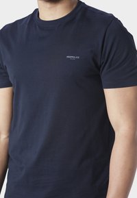 Μπλε σκούρο κοντομάνικο T-shirt με μικρό λευκό λογότυπο «883 Police Milano» στο αριστερό στήθος, φορεμένο από ένα άτομο με το πρόσωπο κομμένο στη μέση.