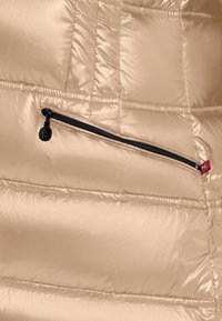 Beżowa kurtka puchowa z połyskującym wykończeniem, z czarną kieszenią na zamek błyskawiczny i czerwoną metką. Szyte pionowe, pikowane sekcje.