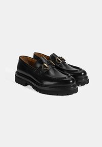 Zwarte leren loafers met een ronde neus, decoratieve gouden hardware, een textuurband en een dikke zwarte rubberen zool met profiel.