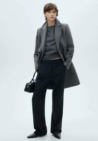 Gray double-breasted coat over a gray sweater, paired with dark pinstriped trousers. Accessories include a small black handbag and pointed shoes.