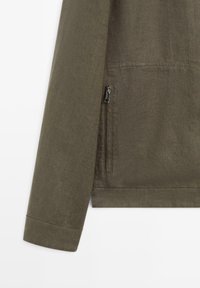 Ærme og nederste side af olivengrøn jakke med synlig lynlåslomme og syning ved manchetter på ensfarvet baggrund.