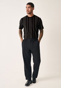 Schwarzer Strickpullover mit kurzen Ärmeln und weißen vertikalen Streifen, kombiniert mit dunkler Hose und schwarzen Schuhen. Glatter Stoff und taillierte Passform.