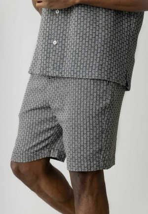 Férfi, aki szürke mintás rövidnadrágot és gombos inget visel, karját keresztbe téve, a comb közepéig és a törzsig vágva, egyszínű háttér előtt.