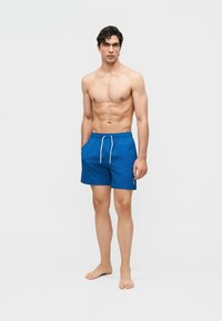 Mężczyzna bez koszuli, stojący boso, ubrany w niebieskie kąpielówki z troczkiem na tle jednolitego jasnego tła.