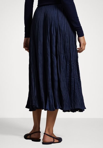 Saia midi plissada na cor navy, com textura enrugada, bainha ampla e uma cintura confortável, combinada com sandálias planas escuras.