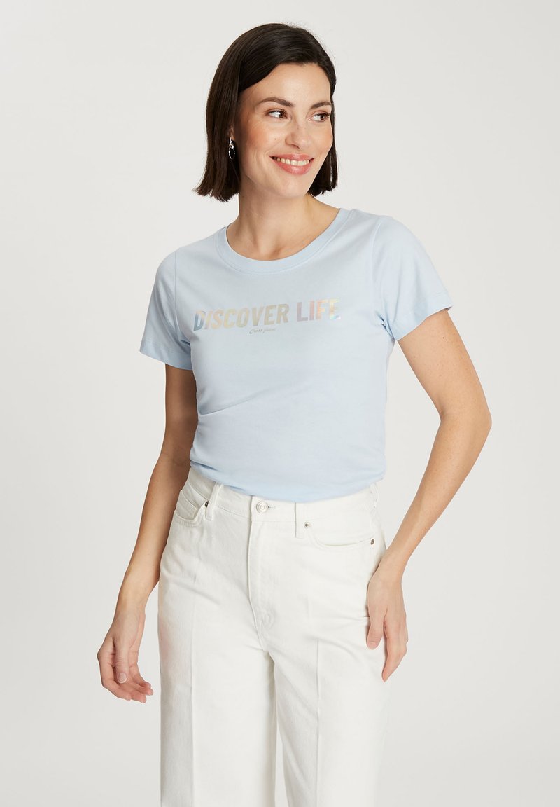 Jasnoniebieska bawełniana koszulka z krótkim rękawem z napisem "ODKRYJ ŻYCIE" w iryzujących literach, zestawiona z białymi spodniami o wysokim stanie.