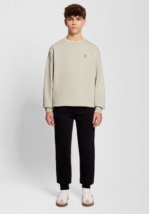 Jonge man die recht naar voren kijkt, gekleed in een beige sweatshirt, zwarte broek en witte sneakers, tegen een eenvoudige lichte achtergrond.