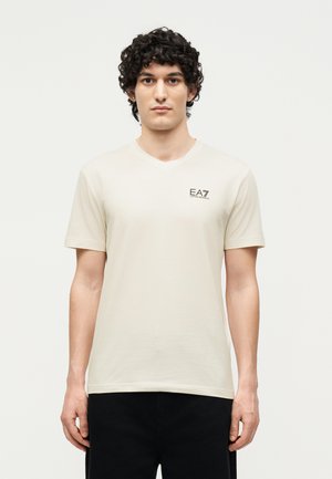 Jeune homme aux cheveux bouclés portant un t-shirt beige clair EA7 Emporio Armani et un pantalon noir, debout devant un fond blanc uni.