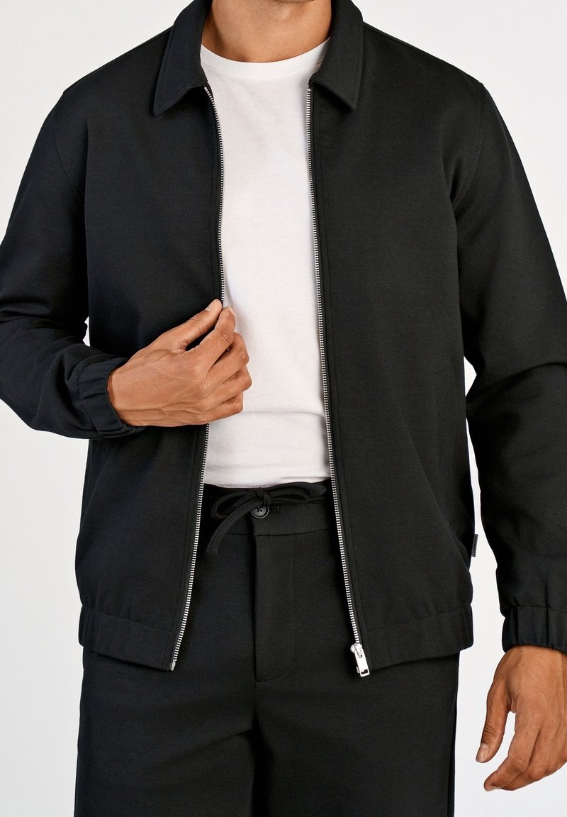 Homme portant une veste noire zippée par-dessus un t-shirt blanc et un pantalon noir, tenant la fermeture éclair de la veste d'une main.