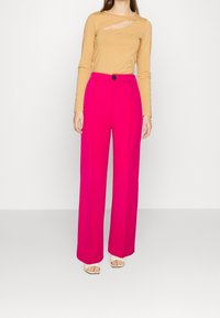 Haut à manches longues en découpe de couleur beige, associé à un pantalon large rose avec une fermeture à boutons sur le devant. Le mannequin porte des sandales neutres.