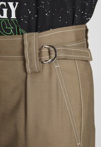 Gros plan sur un pantalon beige avec des coutures blanches, doté d'une boucle de ceinture double anneau métallique et d'une partie d'une chemise noire tachetée avec un texte blanc et vert.