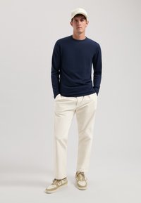 Marineblaue Langarmshirt, cremefarbene Hose, beige Sneakers. Modell trägt eine hellfarbige Mütze. Einfaches Design, glattes Material, lockere Passform.