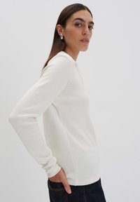 Weißer Pullover mit langen Ärmeln, tailliertem Schnitt, geripptem Muster und Rundhalsausschnitt, kombiniert mit dunklen Jeans. Minimalistischer Stil, leichter Stoff.
