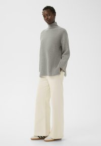 Person wearing a gray ribbed turtleneck sweater, cream wide-leg pants, and white sneakers with black stripes, standing against a plain background.