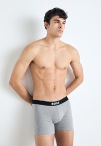 Gray boxer briefs with a black elastic waistband featuring the text "BOSS" in white. Smooth texture and fitted design. Model stands against a plain background.