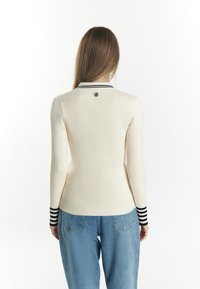 Geribbelde crèmekleurige trui met een aansluitend ontwerp, contrasterende marineblauwe gestreepte kraag en manchetten, met een klein logo op de achterkant. Blauwe baggy jeans.
