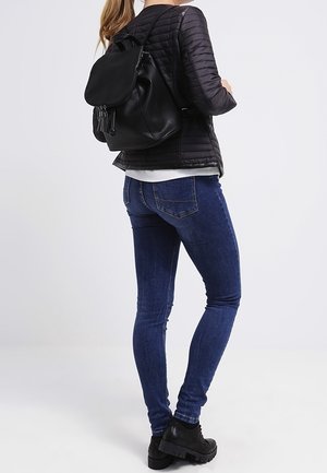 Žena v černé prošívané bundě, modrých úzkých džínách, černých botách a s černým koženým batohem, stojící před jednobarevným pozadím.