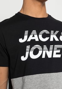 Camiseta de manga corta en negro y gris con letras blancas en negrita "JACK & JONES" con rayas, cuello redondo y una textura suave de algodón.