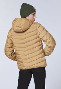 Light brown puffer jacket with a chevron pattern, features a hood, ribbed green beanie, worn with black pants and white side stripes.