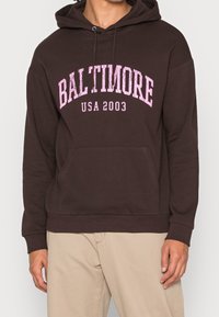 Felpa con cappuccio marrone con la scritta "BALTIMORE USA 2003" in rosa, dotata di tasca a marsupio frontale e polsini e orlo a costine.