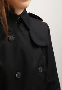 Trench coat negro de doble botonadura con un amplio cuello, botones brillantes y una textura de tela suave. Detalle del cuello ajustable visible.