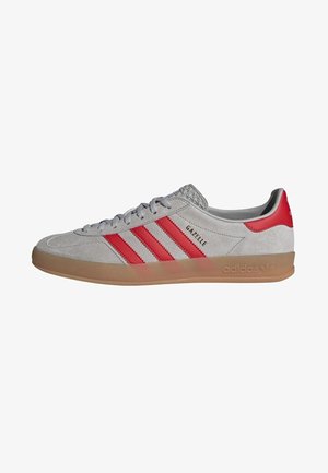 Jasnoszare buty sportowe Adidas Gazelle z czerwonymi paskami, czerwoną taśmą na pięcie, gumową podeszwą i szarymi sznurówkami, widoczne od zewnętrznej strony.