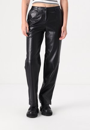 Nainen pukeutunut korkeavyötäröisiin mustiin tekonahkahousuihin, joissa on vyölenkit, yhdistettynä mustiin paksupohjaisiin kenkiin ja lyhyt raidallinen yläosa.