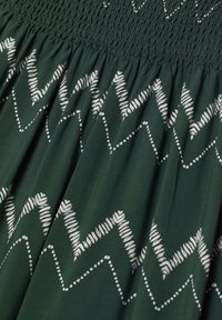 Green fabric dress with smocked bodice, featuring white geometric patterns and accents. The texture is soft, with a flowing design.