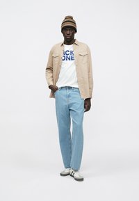 Beige overhemd met knopen over een wit T-shirt met blauwe tekst, lichtblauwe jeans, grijs-witte sneakers en een gebreide muts met patroon.